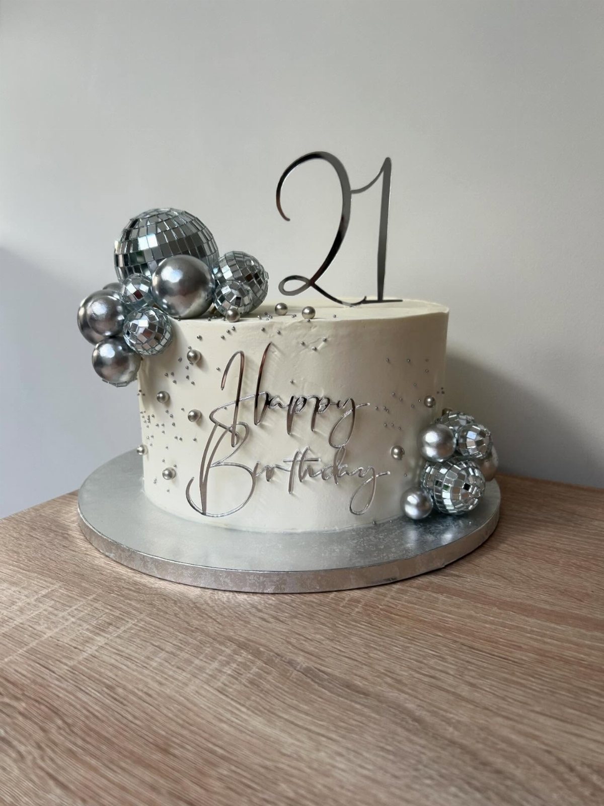 White birthday cake decorated with silver disco balls, metallic spheres, and a black number 21 topper.