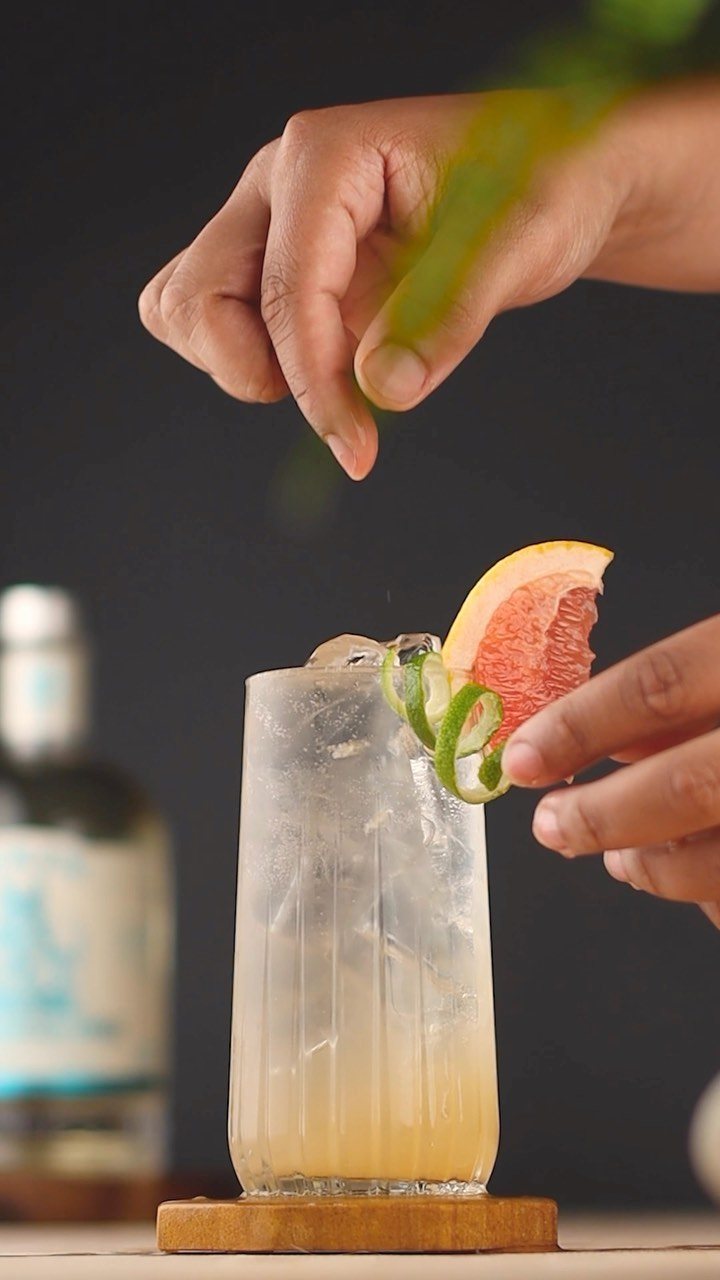 Hands garnishing a pale yellow cocktail with grapefruit and greenery in a ribbed glass.