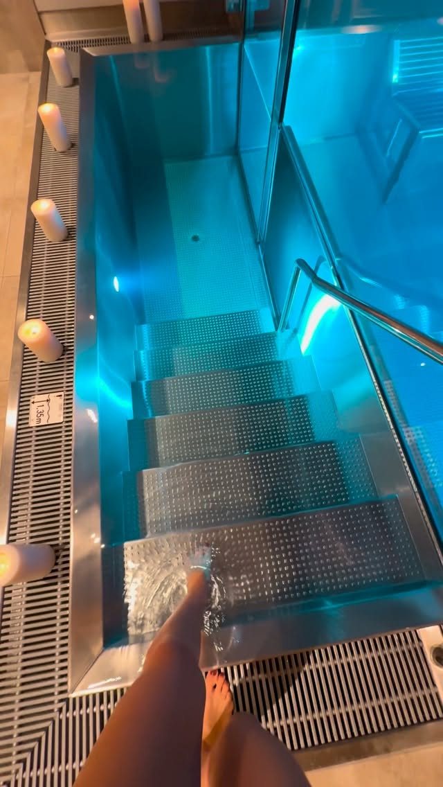 A person dips their feet into a glowing blue indoor spa pool lined with white candles.