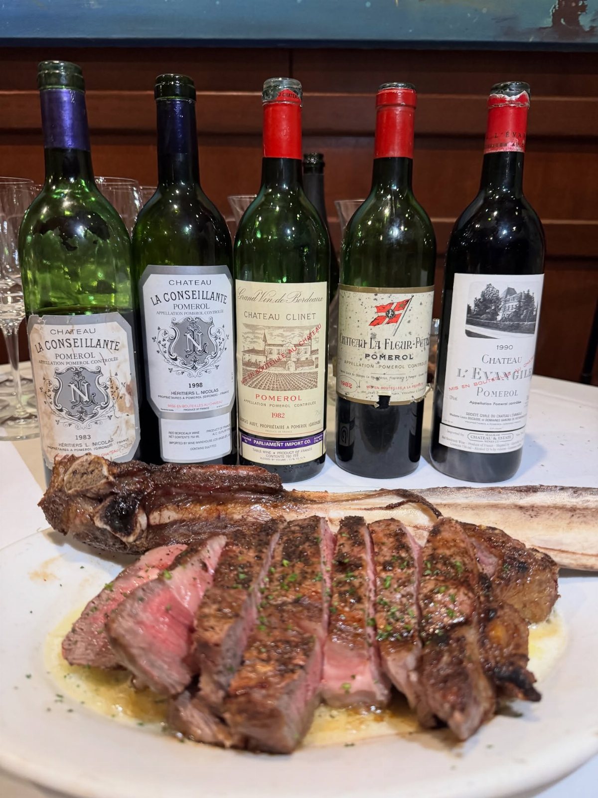 Five wine bottles lined up behind a large plate of sliced steak on a white table.