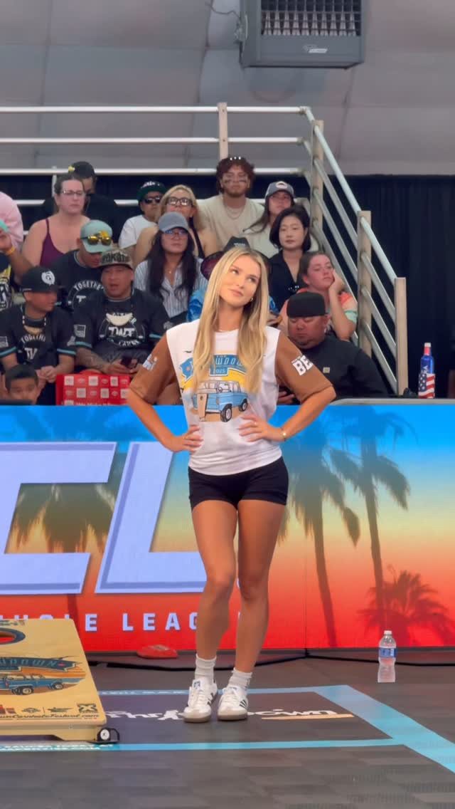 A blonde woman standing in an arena near a cornhole board with spectators in the background.