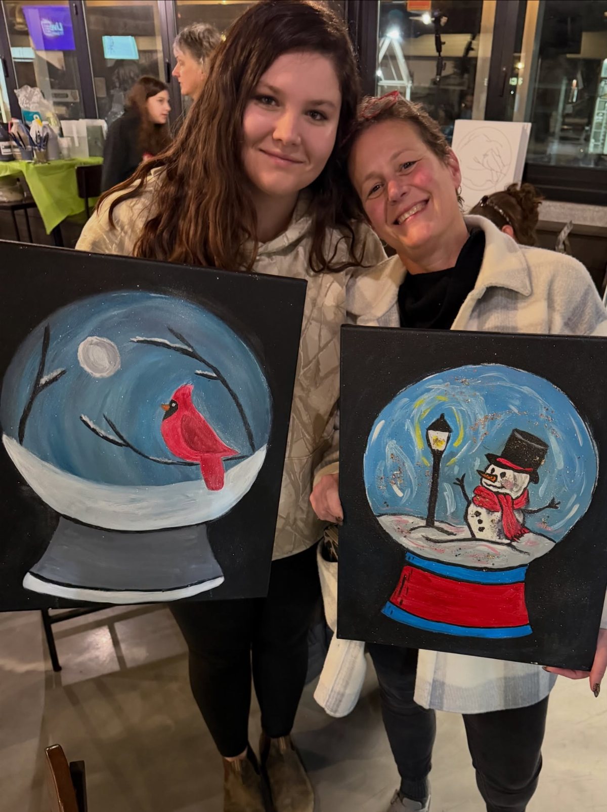 Two women smiling while holding up their completed snow globe themed paintings.