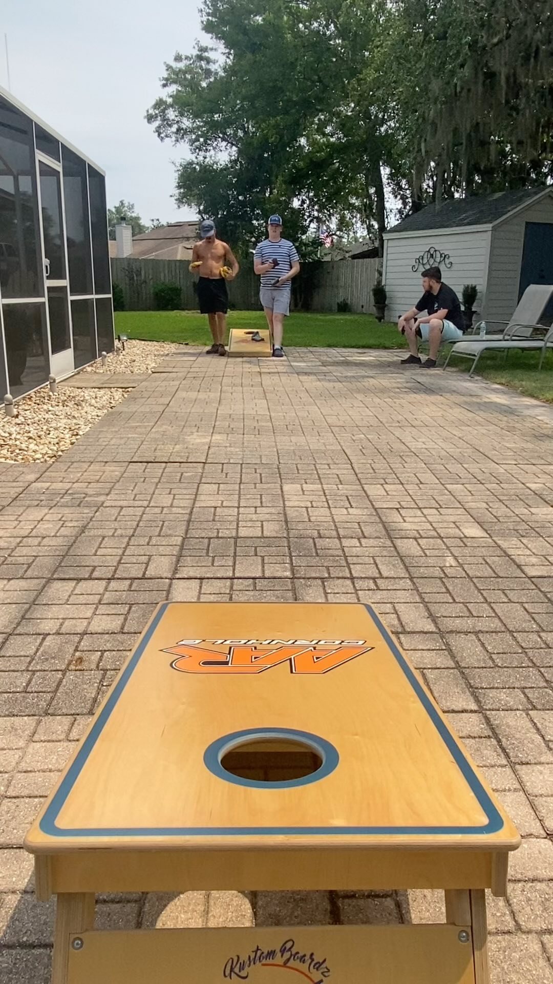 A wooden corn hole board sits on a stone patio with people in the background.
