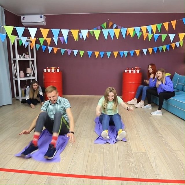 Two people sliding on purple mats across a wooden floor during a party game.
