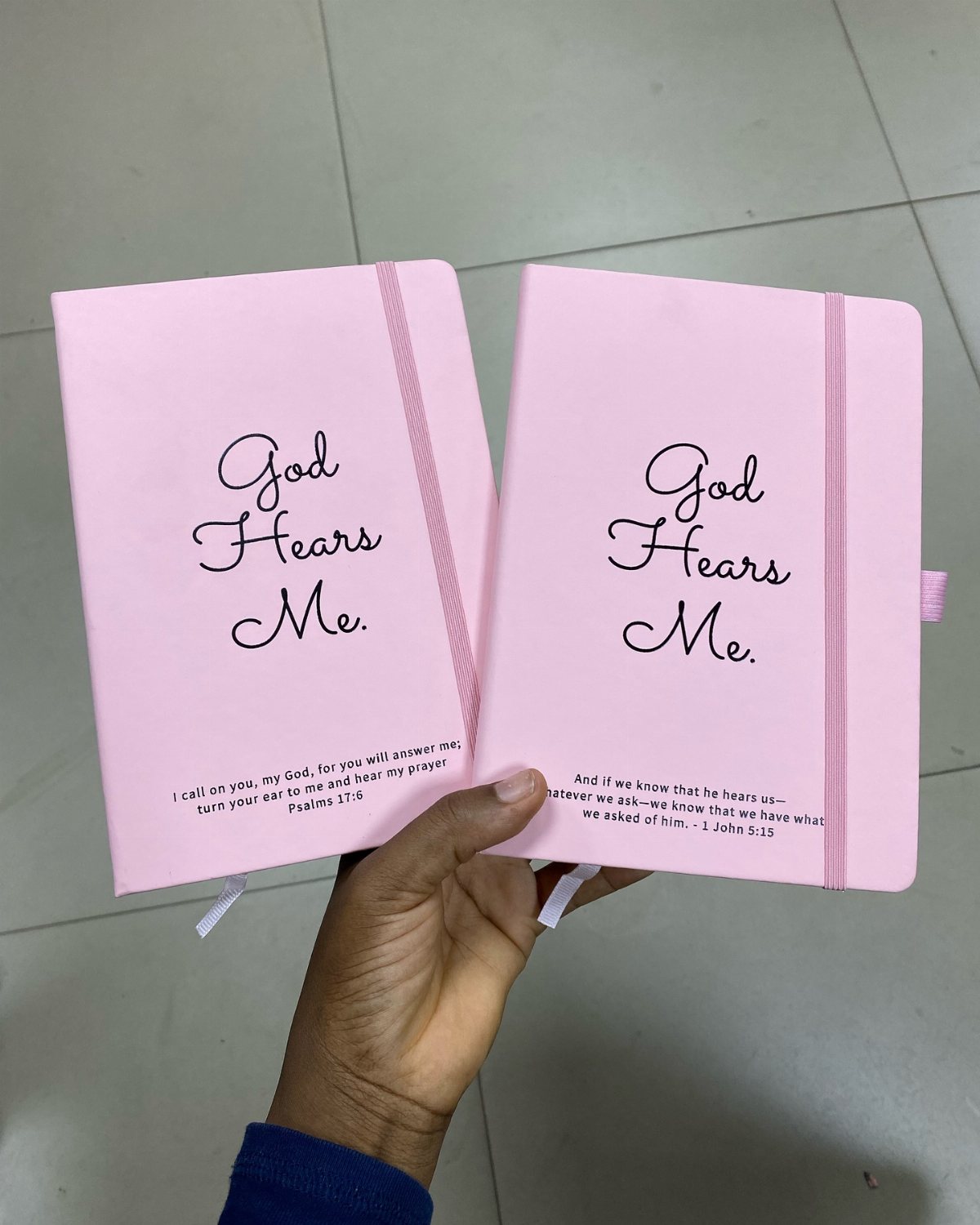 A hand holding two pink journals with the printed text God Hears Me.