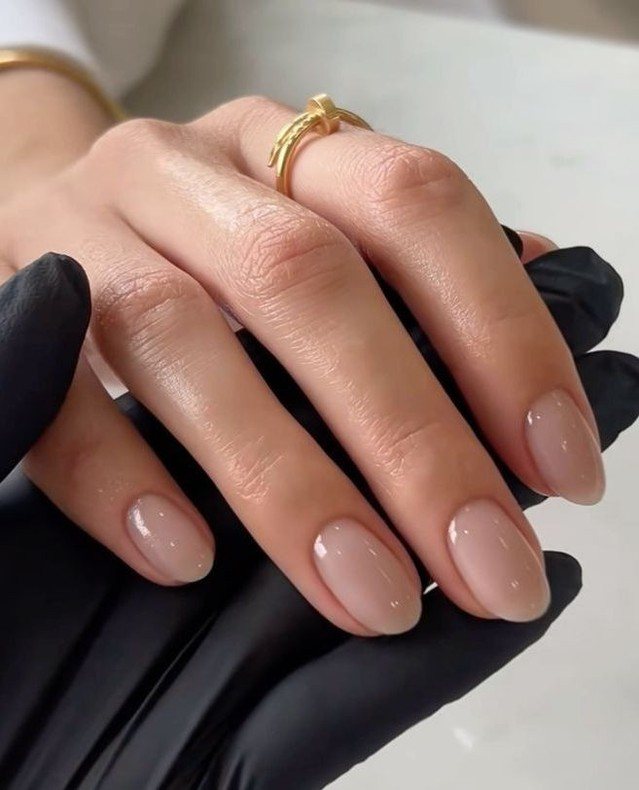 Hands with glossy neutral nude manicured nails and a gold ring.
