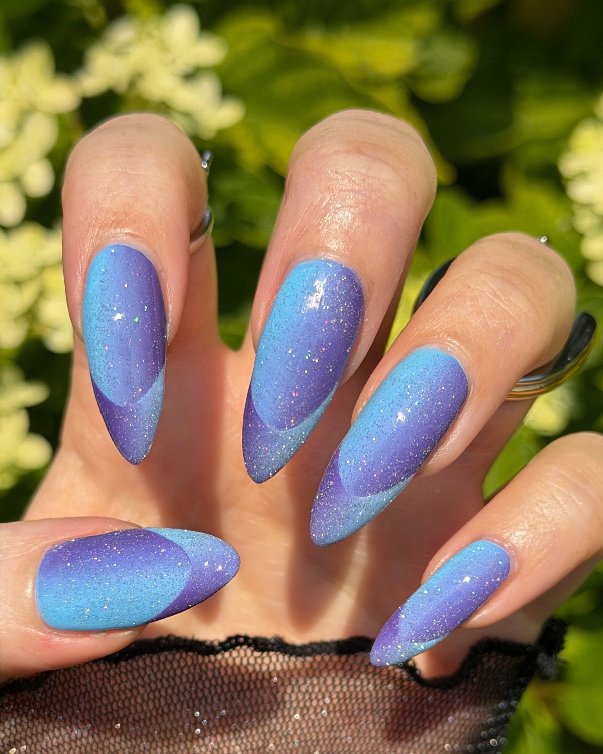 Long almond-shaped nails with blue and purple ombre glitter design against a floral background.