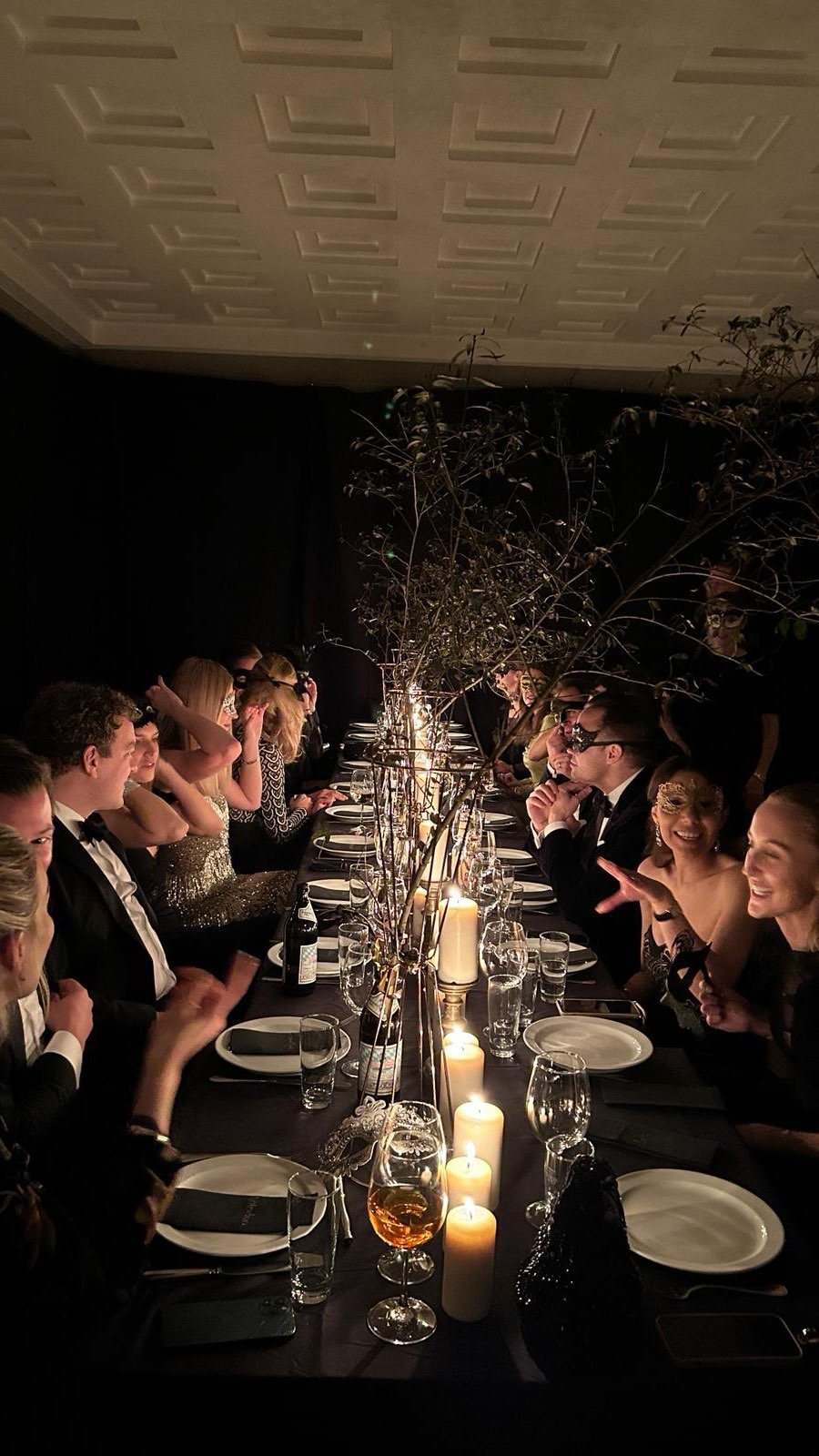 Guests in formal attire sit at a long, candlelit dinner table decorated with branches.