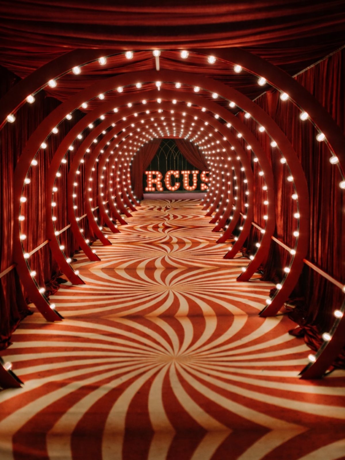 Red and white swirling patterned floor leading through a lighted circular arch tunnel.