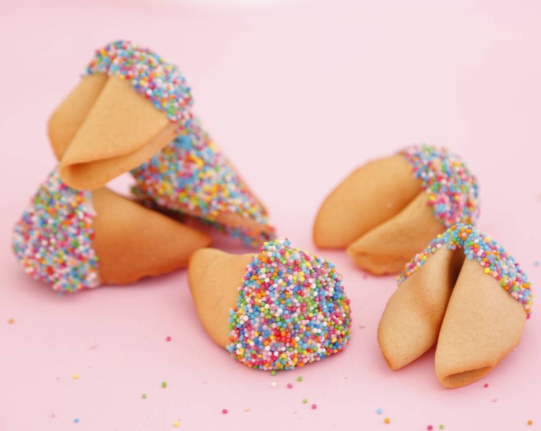 Decorated fortune cookies with colorful rainbow sprinkles scattered on a pink background.