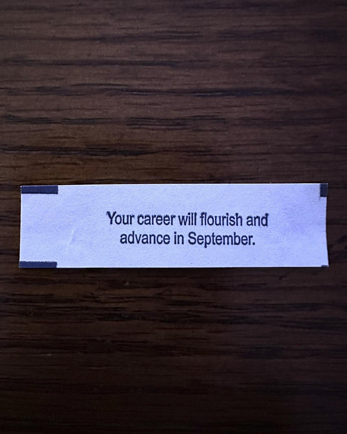 A white paper fortune cookie slip with blue text on a dark wood surface.