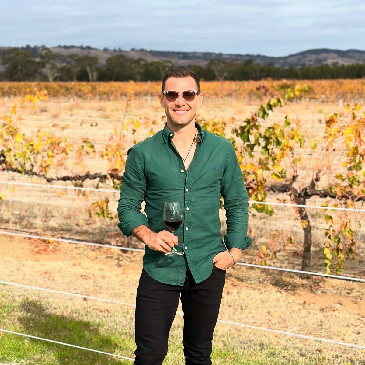 A smiling man in a green shirt holding a glass of red wine in a vineyard.
