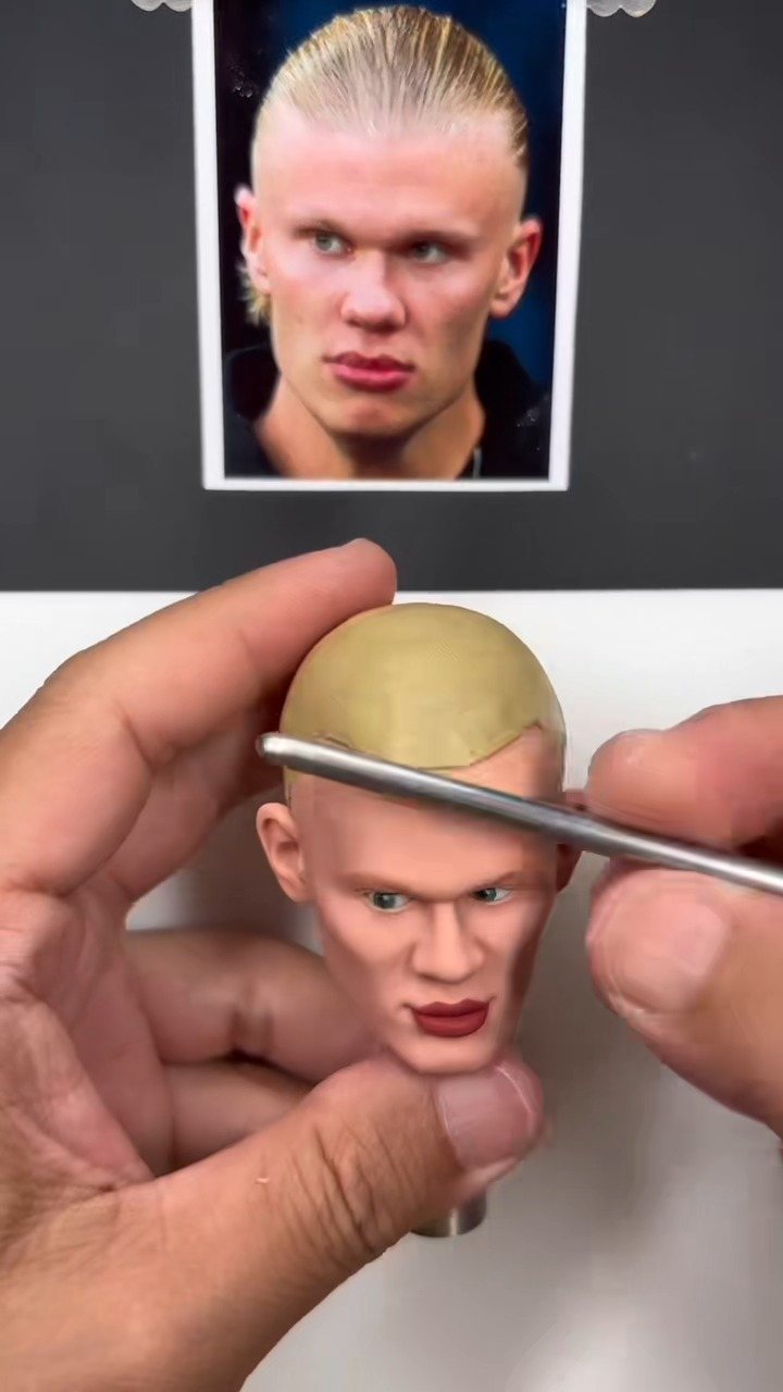 Hands using a metal tool to sculpt a custom bobblehead based on a reference photo.