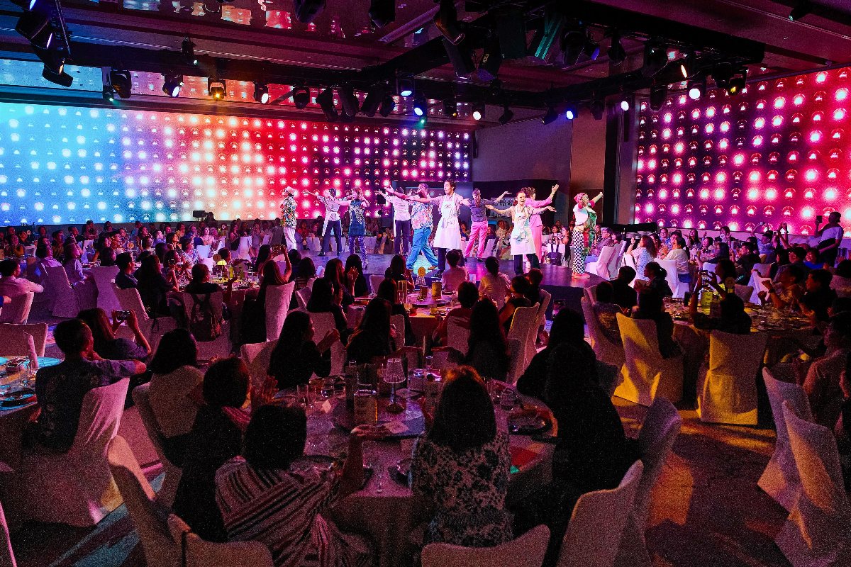Performers dancing on a brightly lit stage before an audience seated at banquet tables.