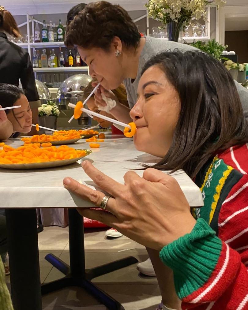 Three women participate in a funny straw-eating game with orange slices at a table.