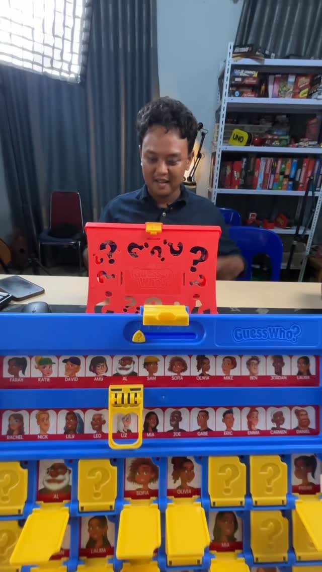 A man sitting behind an open blue and red Guess Who board game on a table.