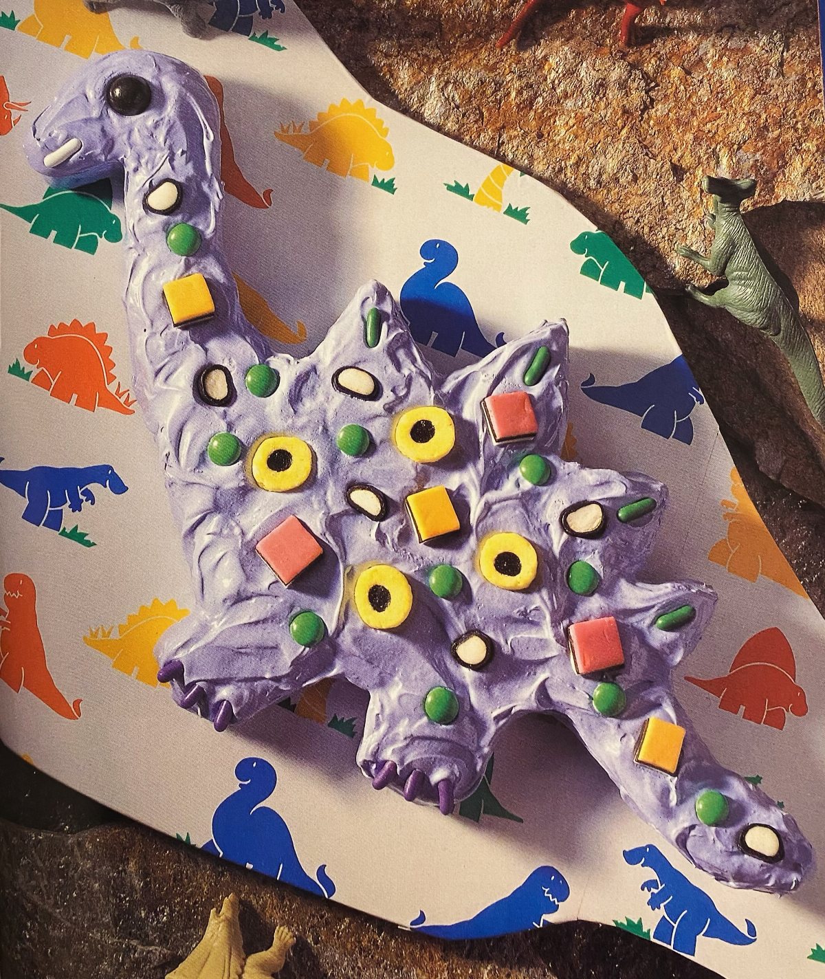 A purple dinosaur-shaped cake decorated with colorful candies on dinosaur patterned paper.