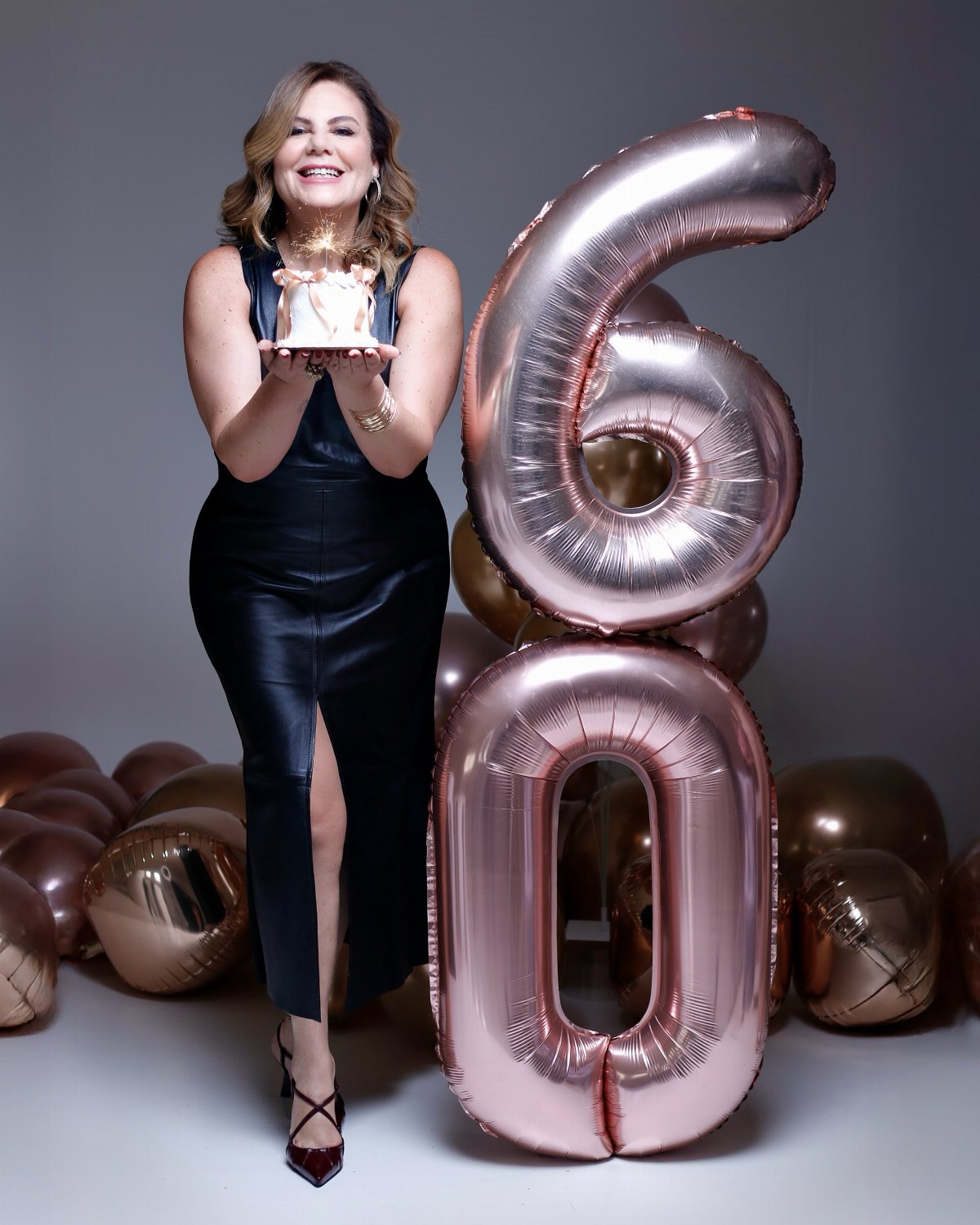 A smiling woman in a black dress holding a cake next to large rose gold 60 balloons.