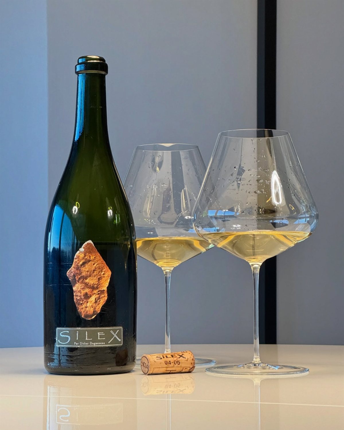 A bottle of Silex wine next to two glasses of white wine on a white surface.