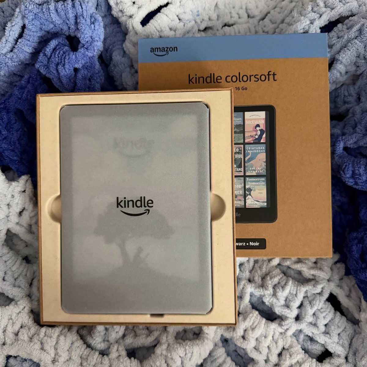 An Amazon Kindle electronic reader in its original packaging resting on a blue and white crochet blanket.