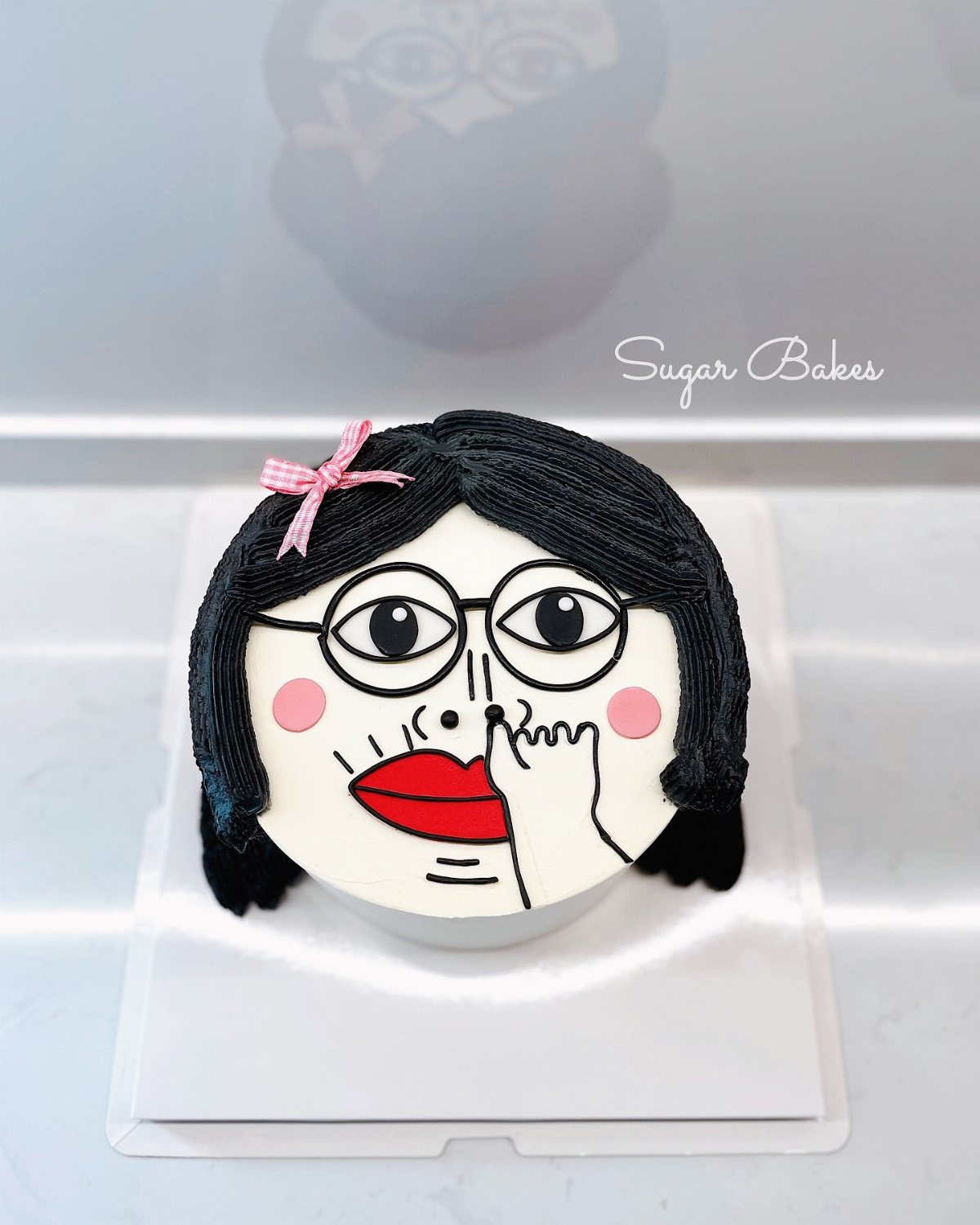 A round white cake decorated with a stylized black-haired female face wearing glasses.