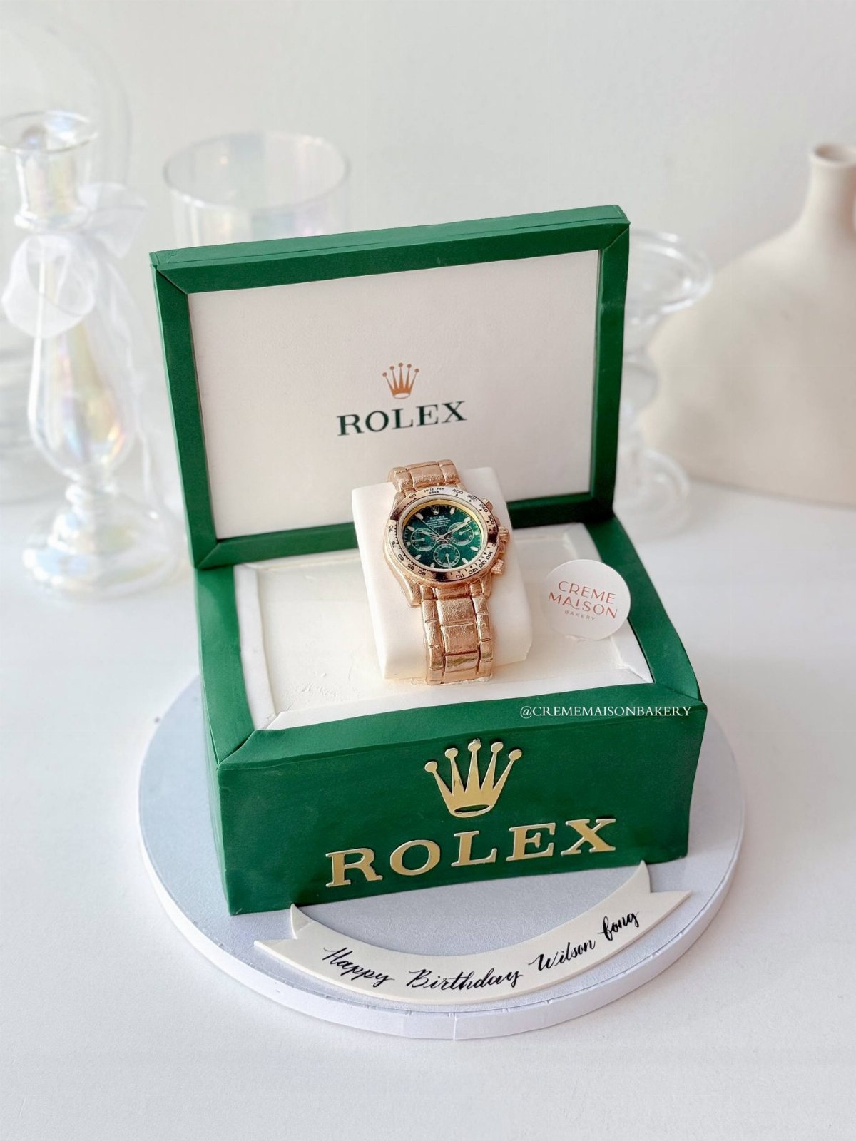 A hyper-realistic cake designed to look like an open green Rolex watch box with an edible gold watch.