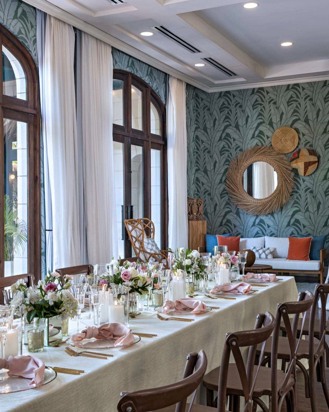 A long dining table set with elegant floral arrangements, pink napkins, and gold cutlery in a tropical room.