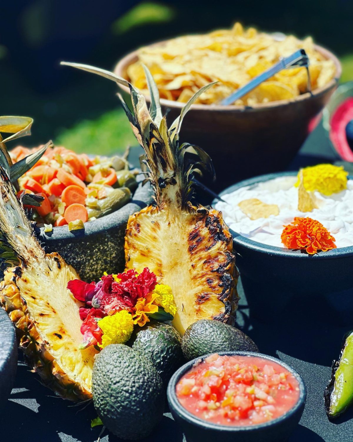 Grilled pineapple, avocados, and salsa bowls arranged on a colorful outdoor food table.