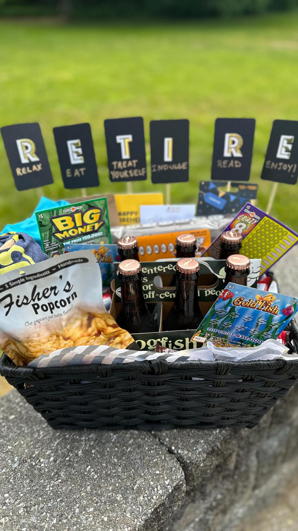 A black wicker basket filled with various snacks and beer bottles against a green lawn.