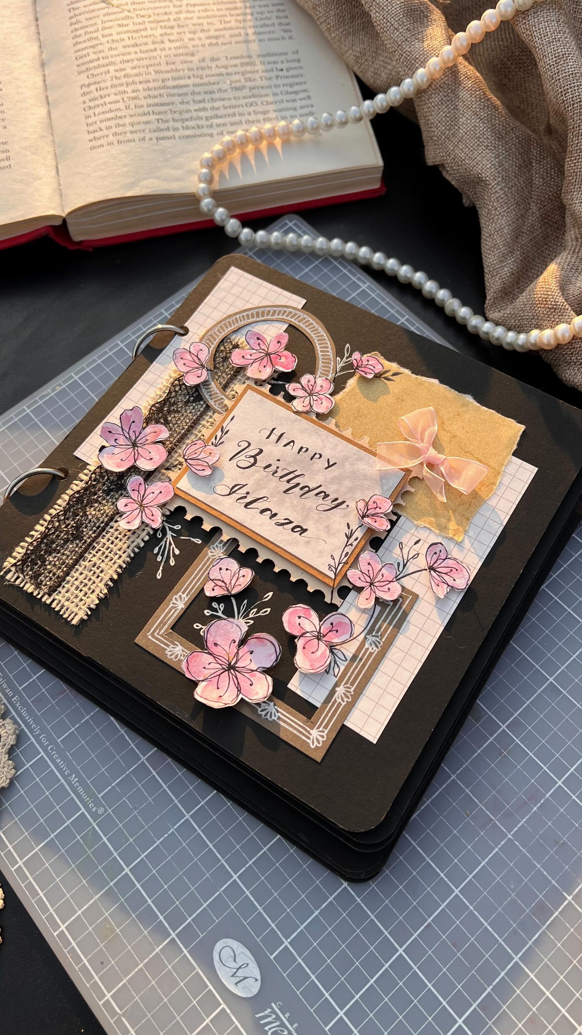 A decorated black scrapbook with pink flowers and a birthday sign on a cutting mat.