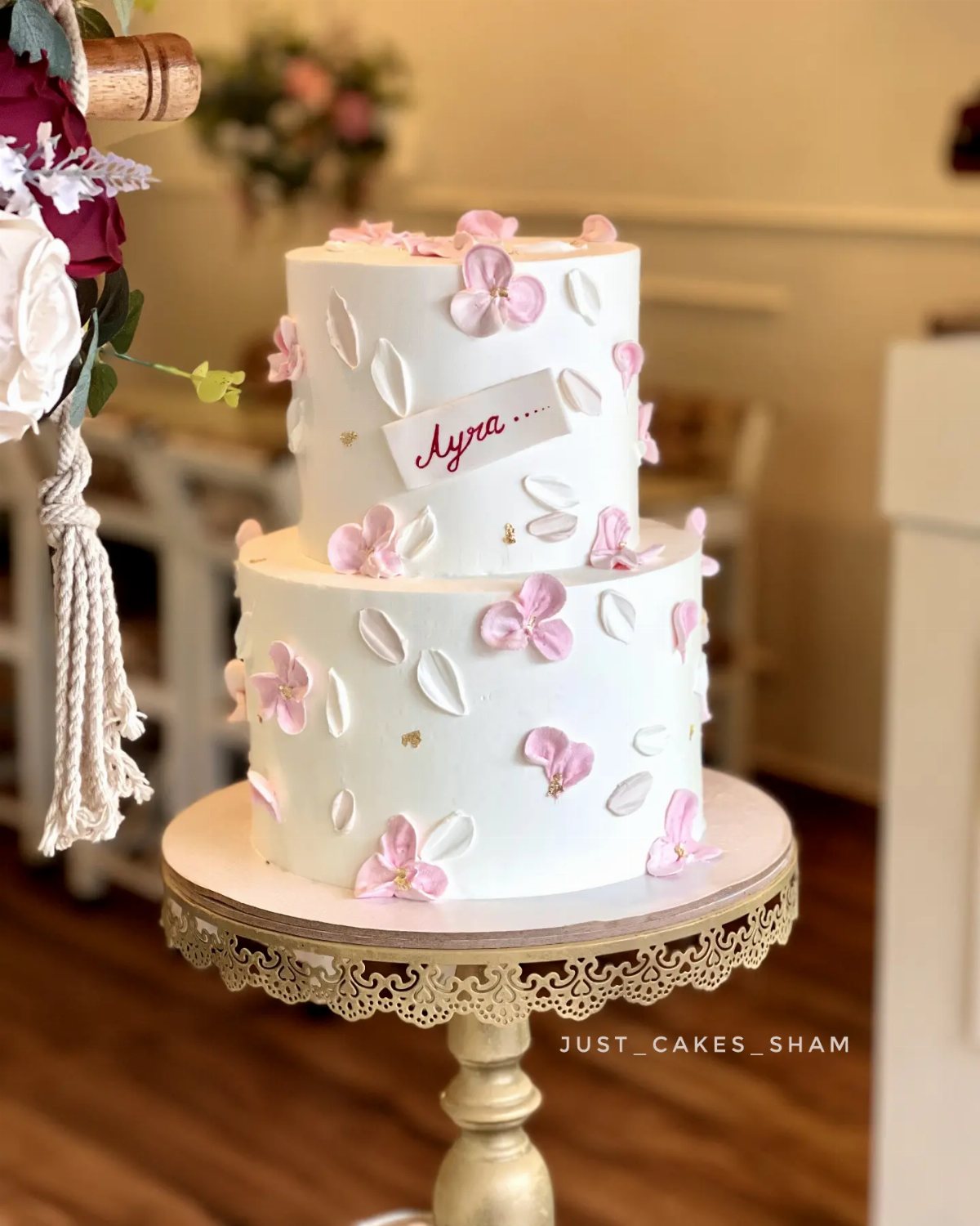 Two-tiered white cake decorated with pink flowers and a name plaque on a gold stand.