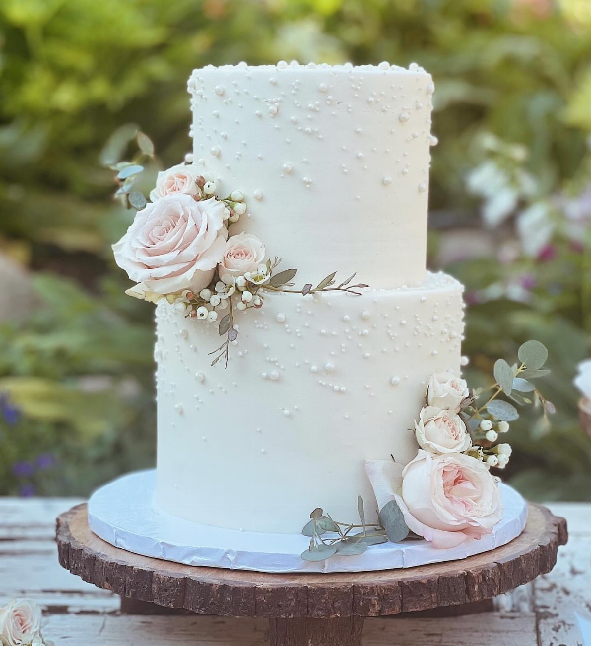 Two-tiered white pearl-decorated wedding cake adorned with pale pink roses and eucalyptus leaves on a wooden stand.
