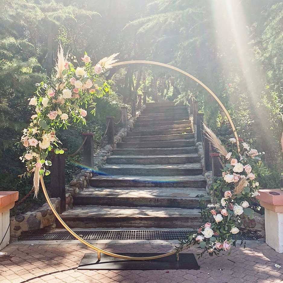 13 Archway for Wedding Ceremony Ideas That Actually Photograph Well 2 Gold circular wedding arch with pink roses positioned before a stone staircase in a forest.
