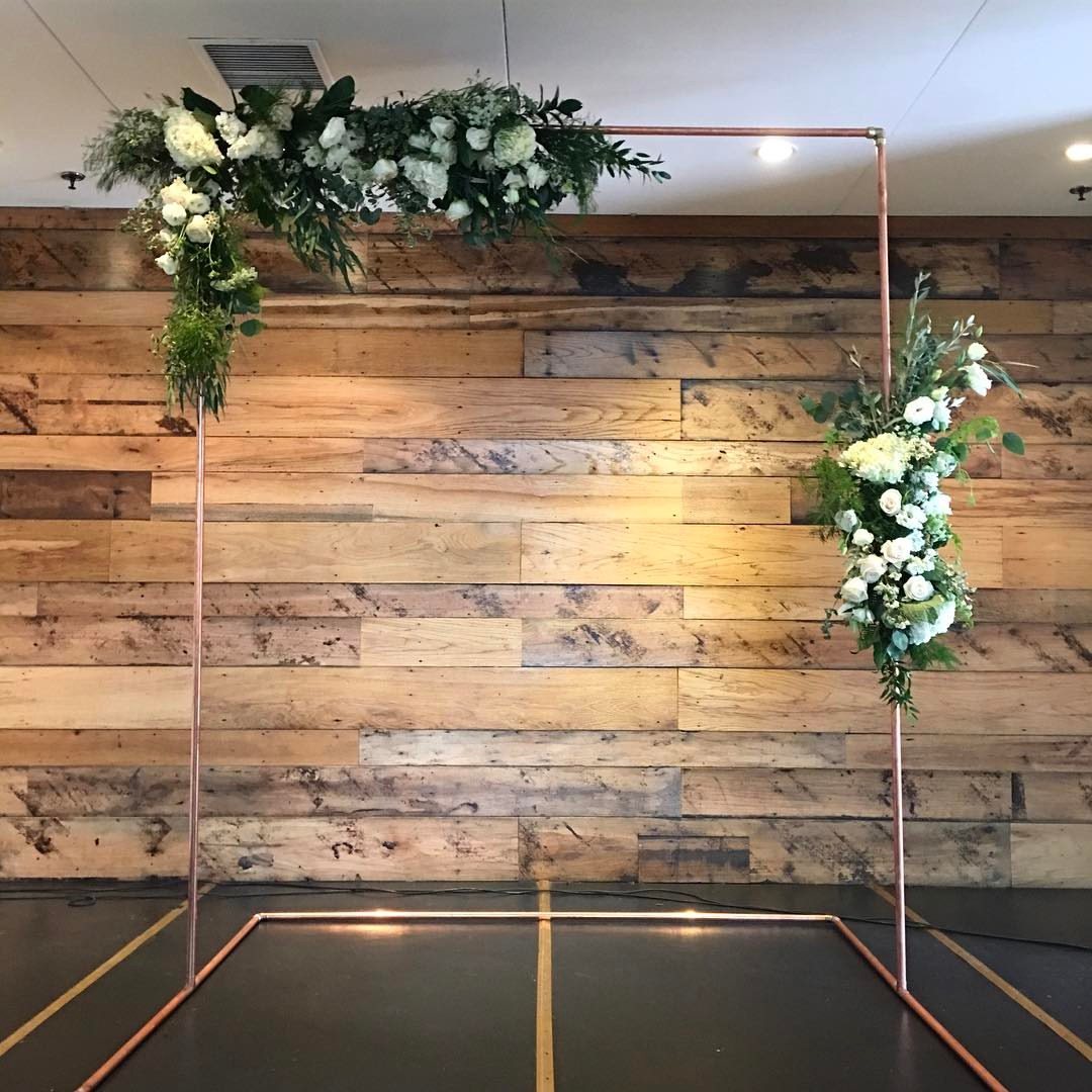 13 Archway for Wedding Ceremony Ideas That Actually Photograph Well 7 Copper pipe wedding arch decorated with white flowers and green foliage against a wooden wall.