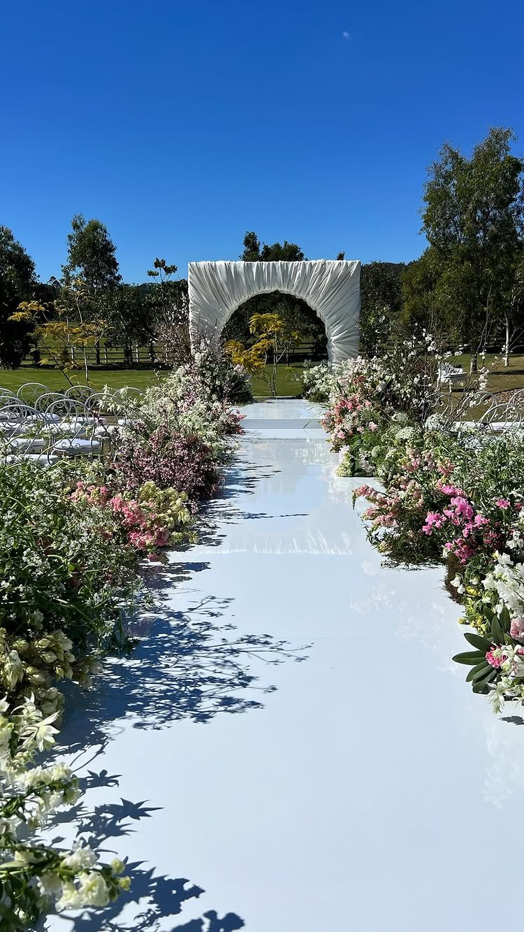 13 Archway for Wedding Ceremony Ideas That Actually Photograph Well 6 White fabric draped over an archway at the end of a floral-lined aisle outdoors.