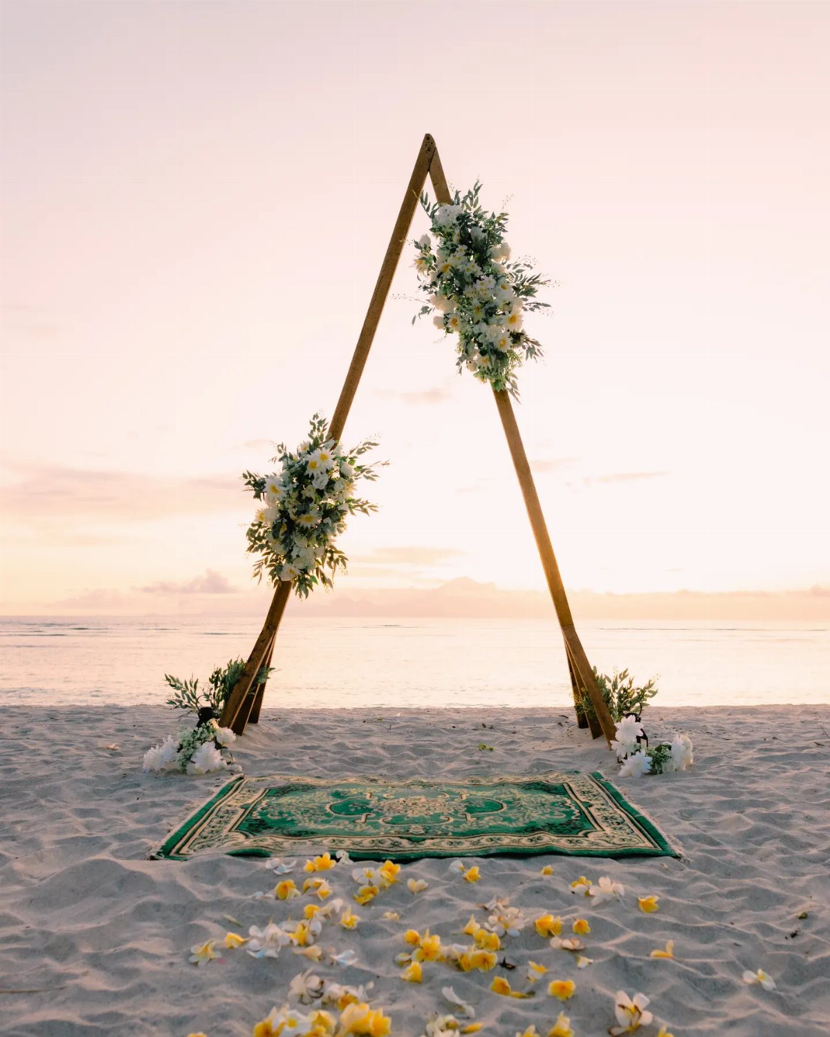 13 Archway for Wedding Ceremony Ideas That Actually Photograph Well 4 A wooden triangle wedding arch with white flowers stands on a beach at sunset.