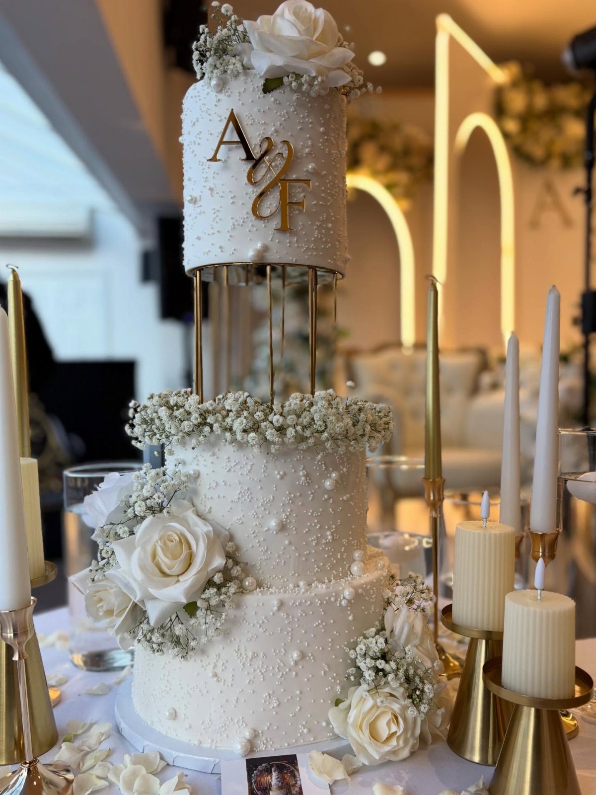 A three-tiered white wedding cake decorated with white roses, baby's breath, and gold monogrammed initials.