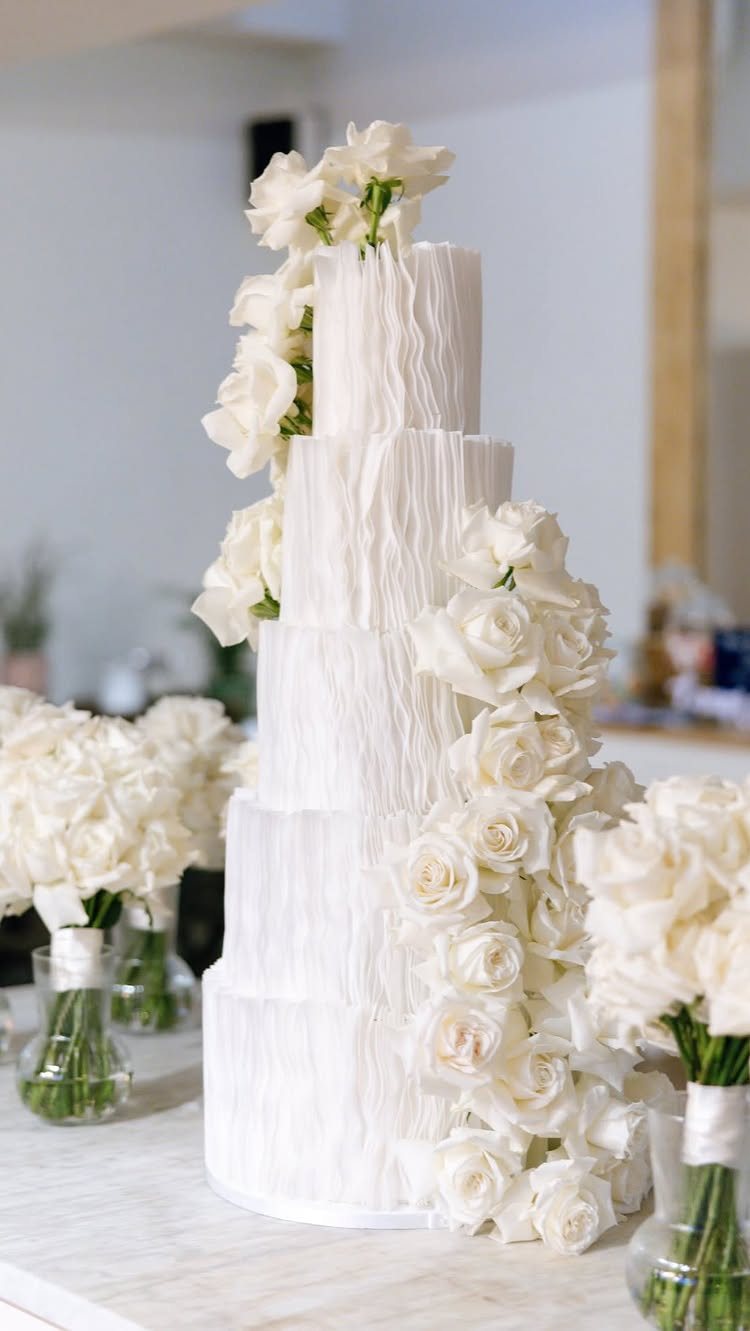 Four-tiered white textured wedding cake decorated with cascading white roses on a white surface.