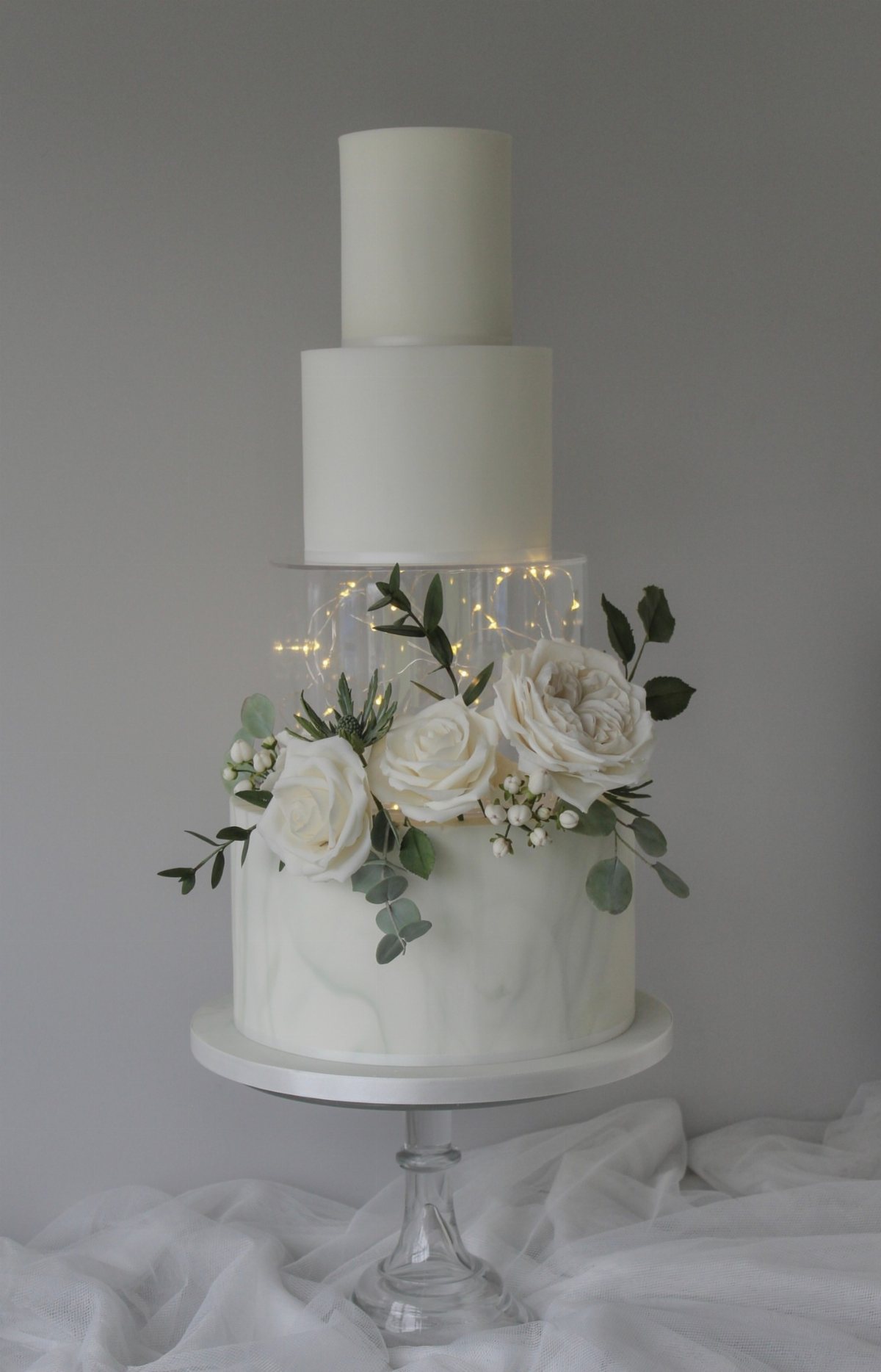 Three-tier white wedding cake featuring fairy lights in a clear middle tier and white rose decorations.