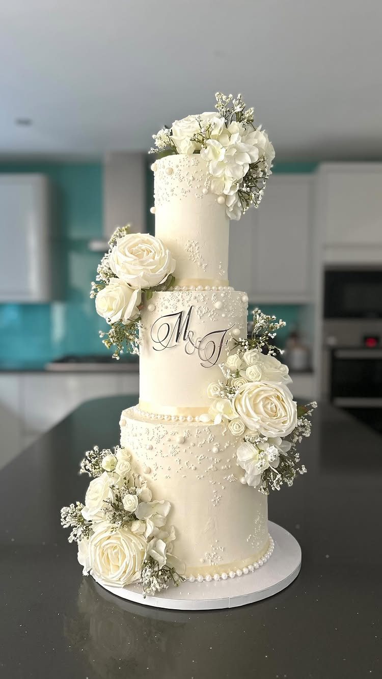 Four-tier white wedding cake decorated with white roses, baby's breath, and elegant initials.