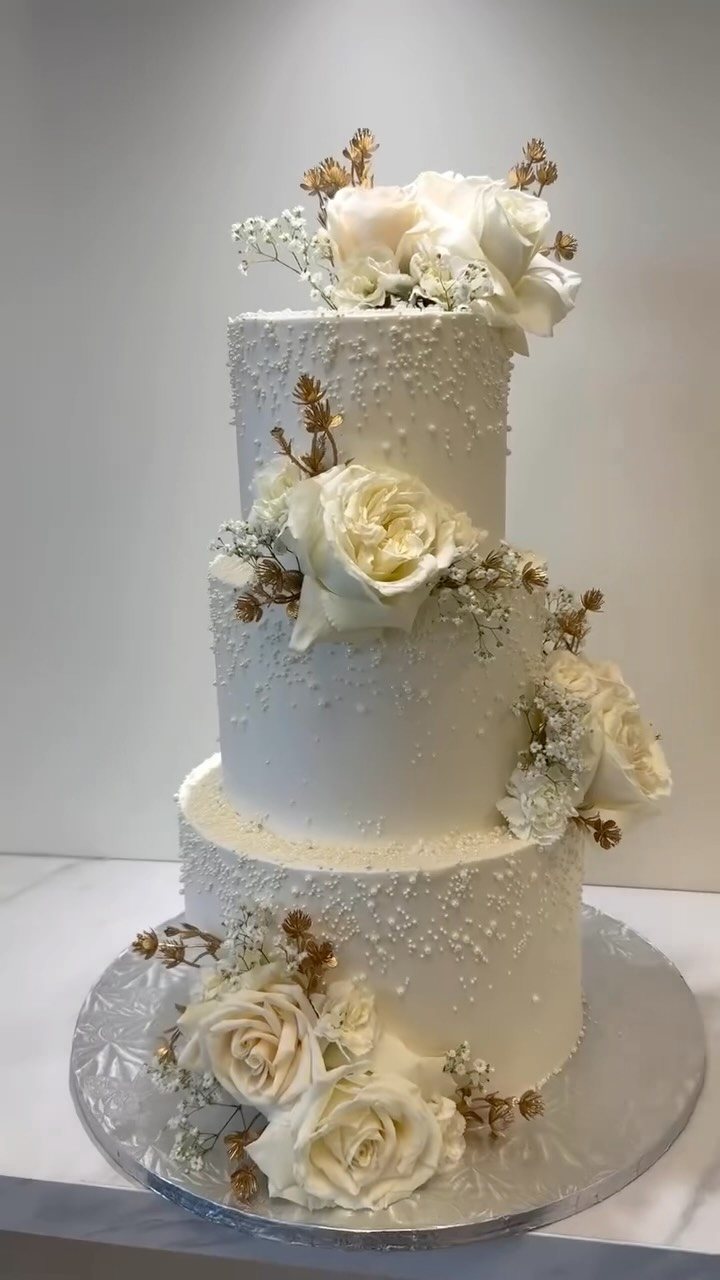 Three-tiered white wedding cake decorated with white roses and delicate pearl frosting details.
