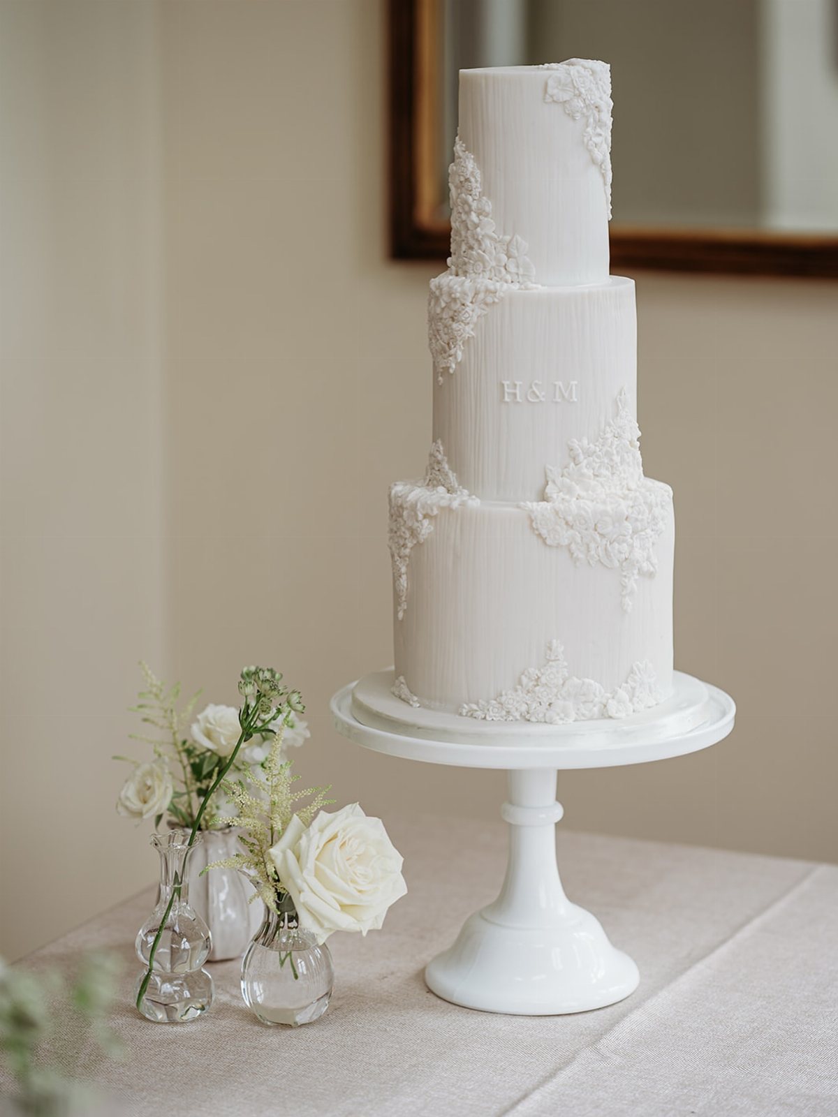 Three-tiered white wedding cake with vertical texture and lace-like sugar floral decorations on a pedestal stand.