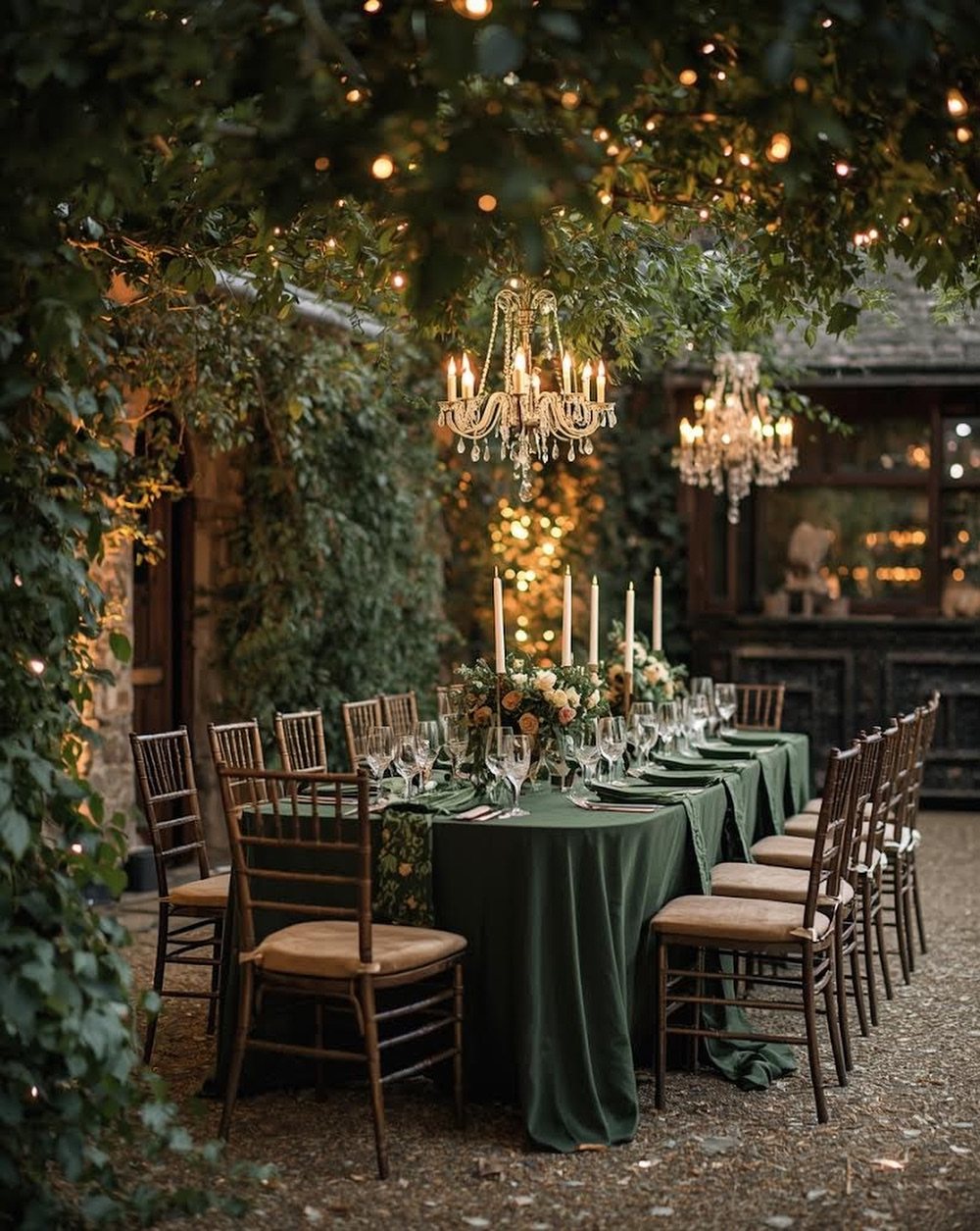 A long dining table set with green linens and candles under hanging chandeliers and trees.