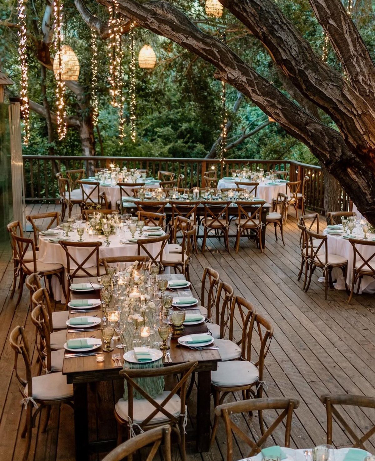 Wedding reception tables set with candles and white linens on a wooden deck under trees.