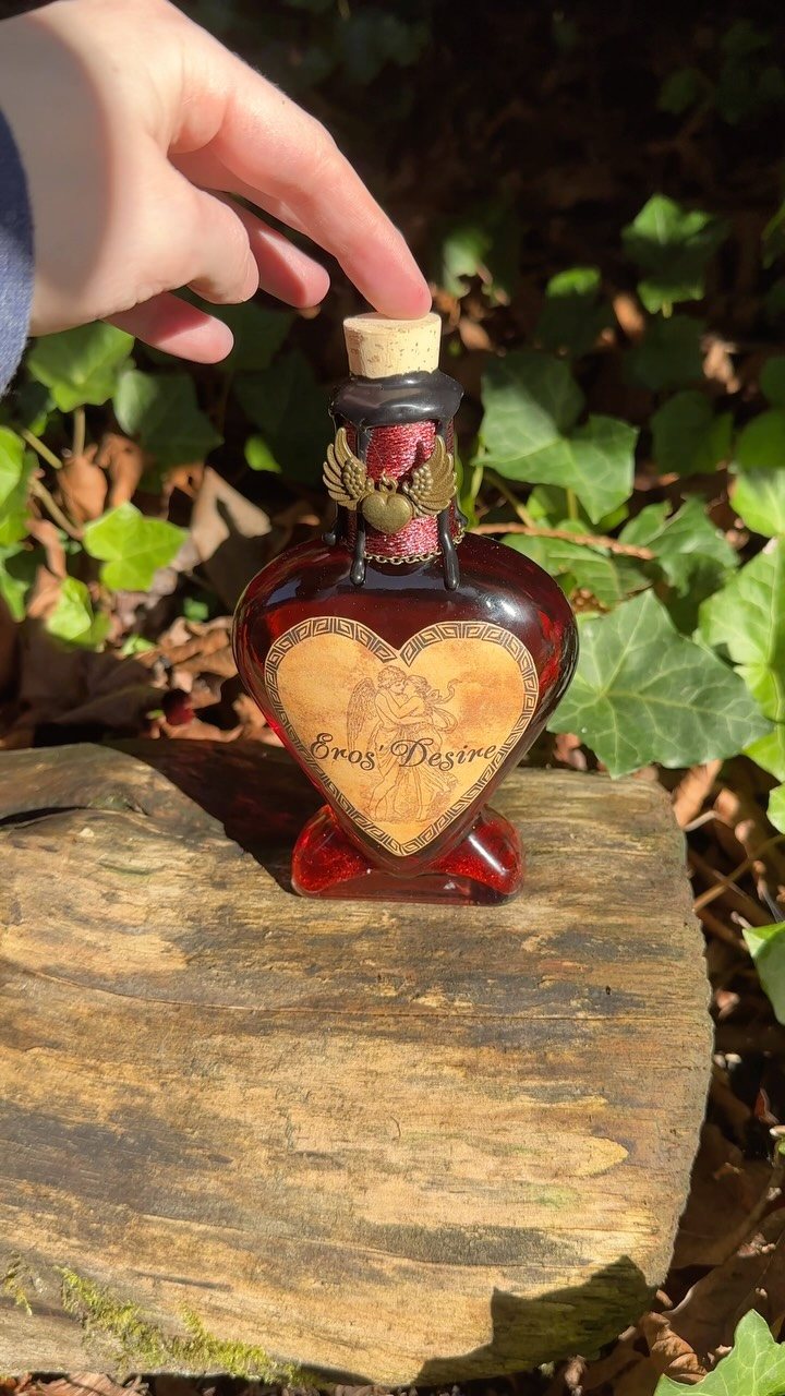 A hand reaches for a heart-shaped red glass bottle labeled Eros Desire on a log.