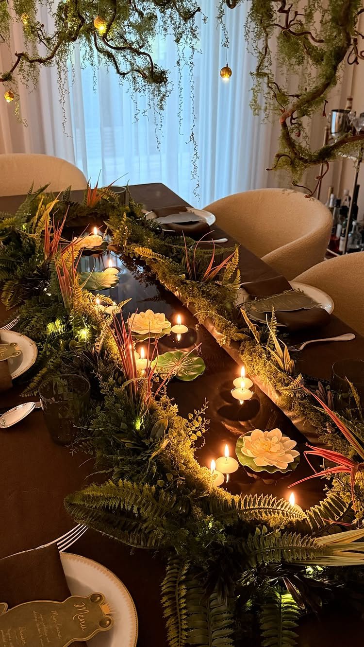 A long dining table decorated with a lush moss and fern centerpiece with glowing candles.