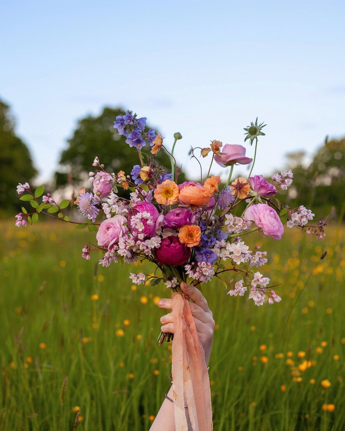 A hand holding a vibrant bouquet of pink, orange, and purple wildflowers in a meadow.