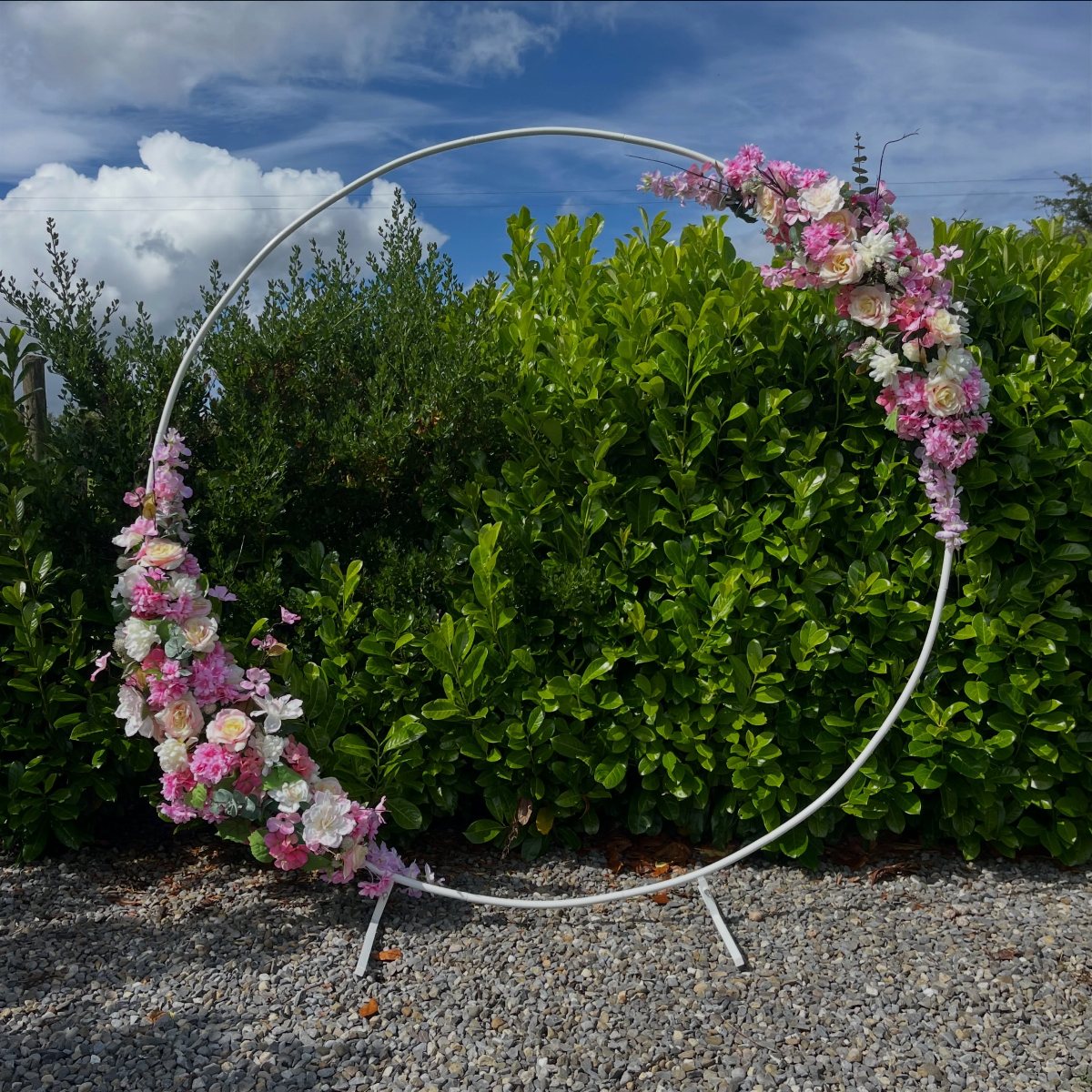 A white circular moon gate arch adorned with pink and white flowers against a green hedge.
