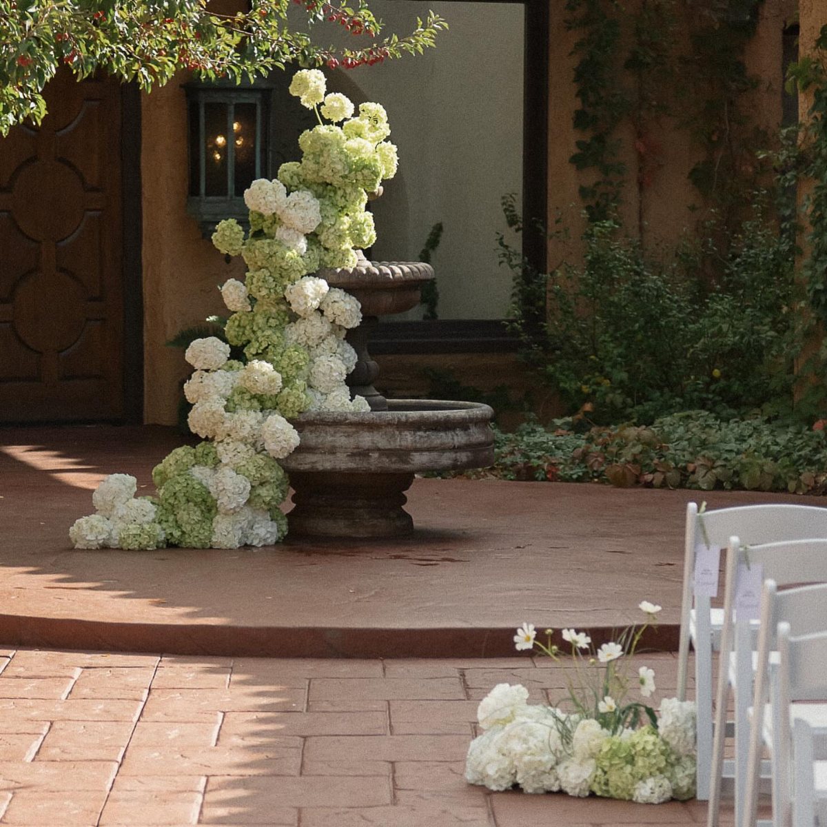White and green hydrangeas cascading down a stone fountain on a brick patio.