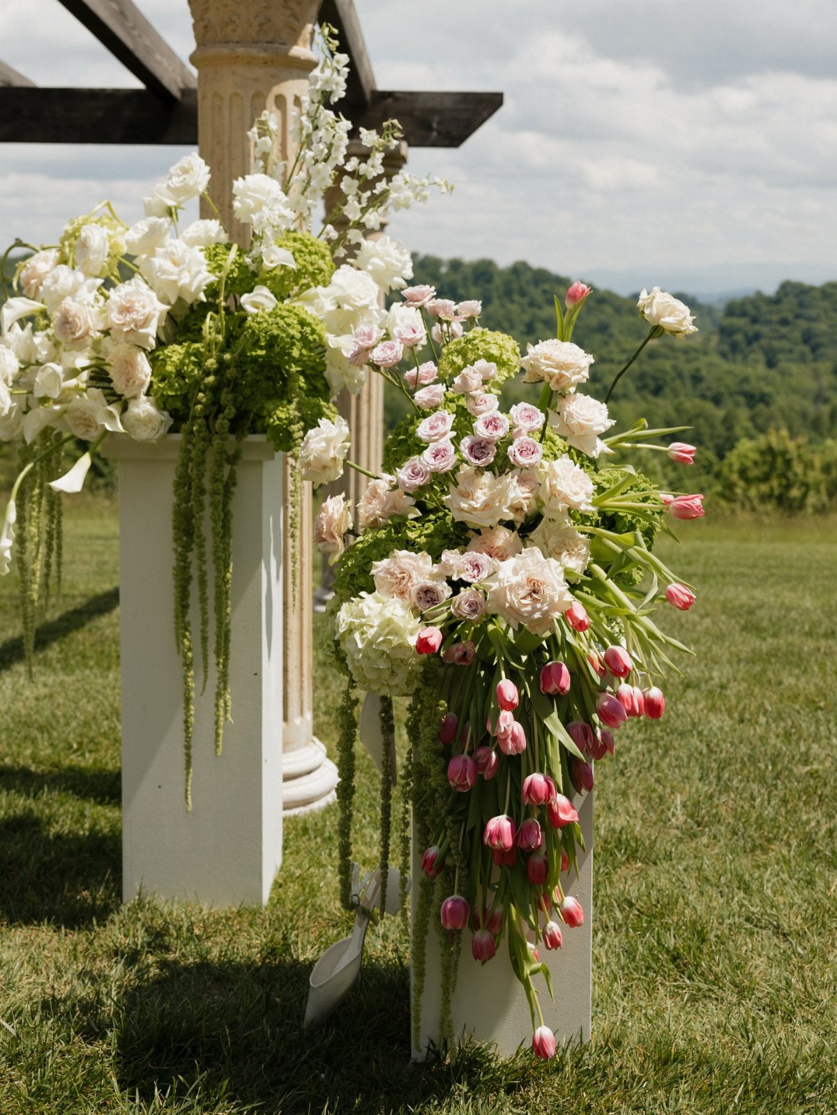 Pink and white floral arrangements with cascading greenery sit on white pedestals in a grassy field.
